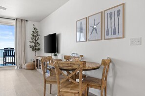 Ein Essbereich mit einem runden Holztisch, vier Holzstühlen, einem an der Wand befestigten Flachbildfernseher und drei gerahmten Kunstwerken mit maritimer Themengebung.