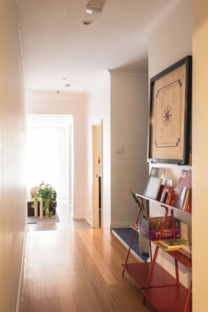 A hallway with a wooden floor, a framed picture on the wall, and a bookshelf with books and a board game. - Flinder's Keepers | Artist's Retreat | 3BR+Parking (Melbourne)