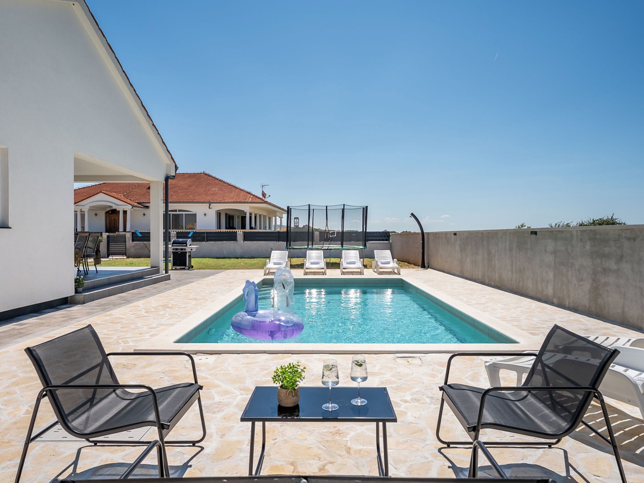 A swimming pool with lounge chairs, a table with drinks, and a view of a house in the background.