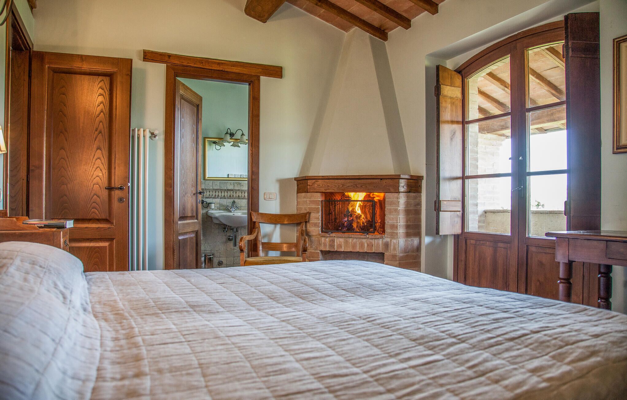 A bedroom with a large bed, a fireplace, wooden doors, and a view of the outside.
