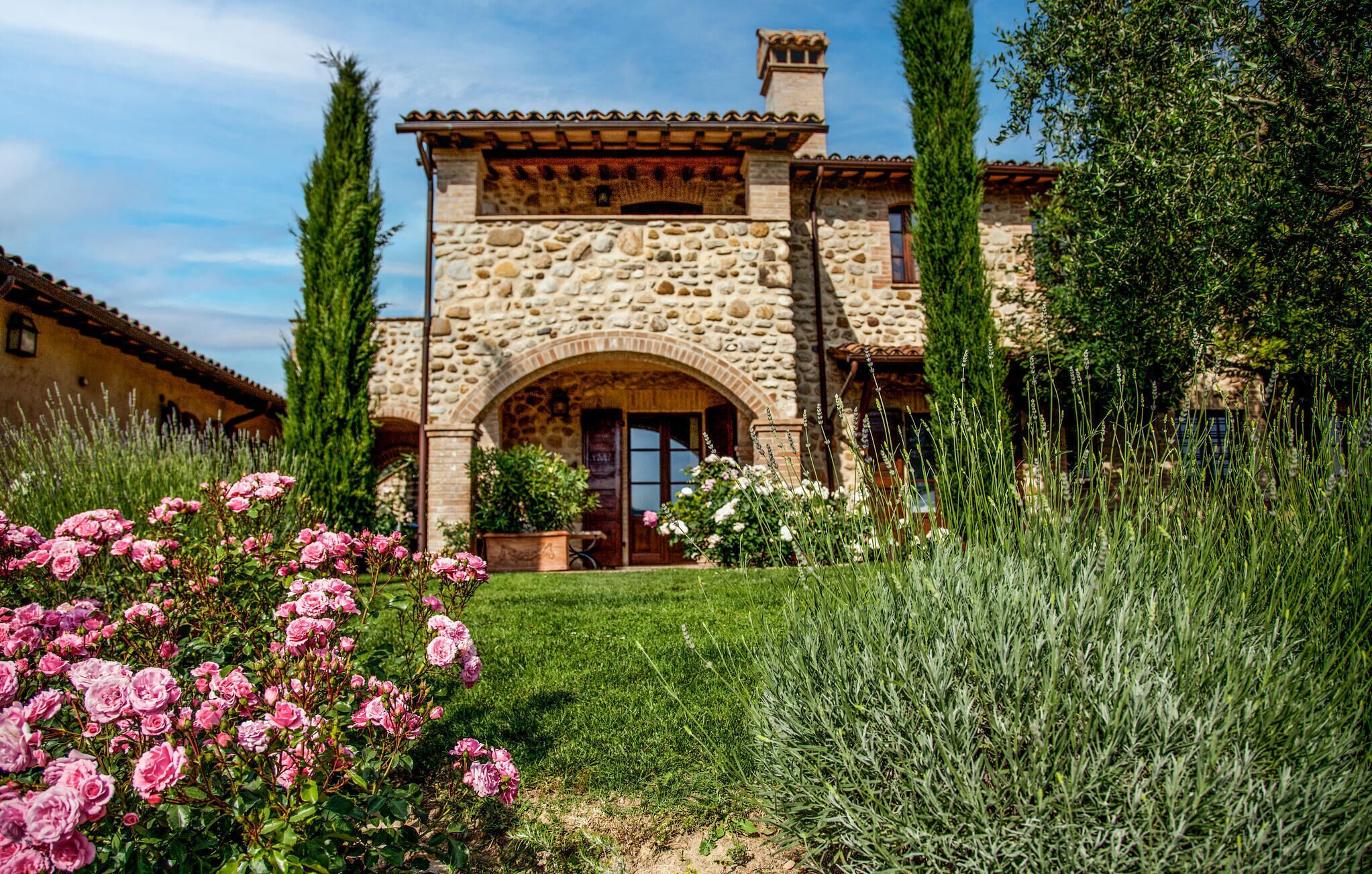 A stone building with a central archway, surrounded by a well-maintained garden with pink roses and tall grasses.