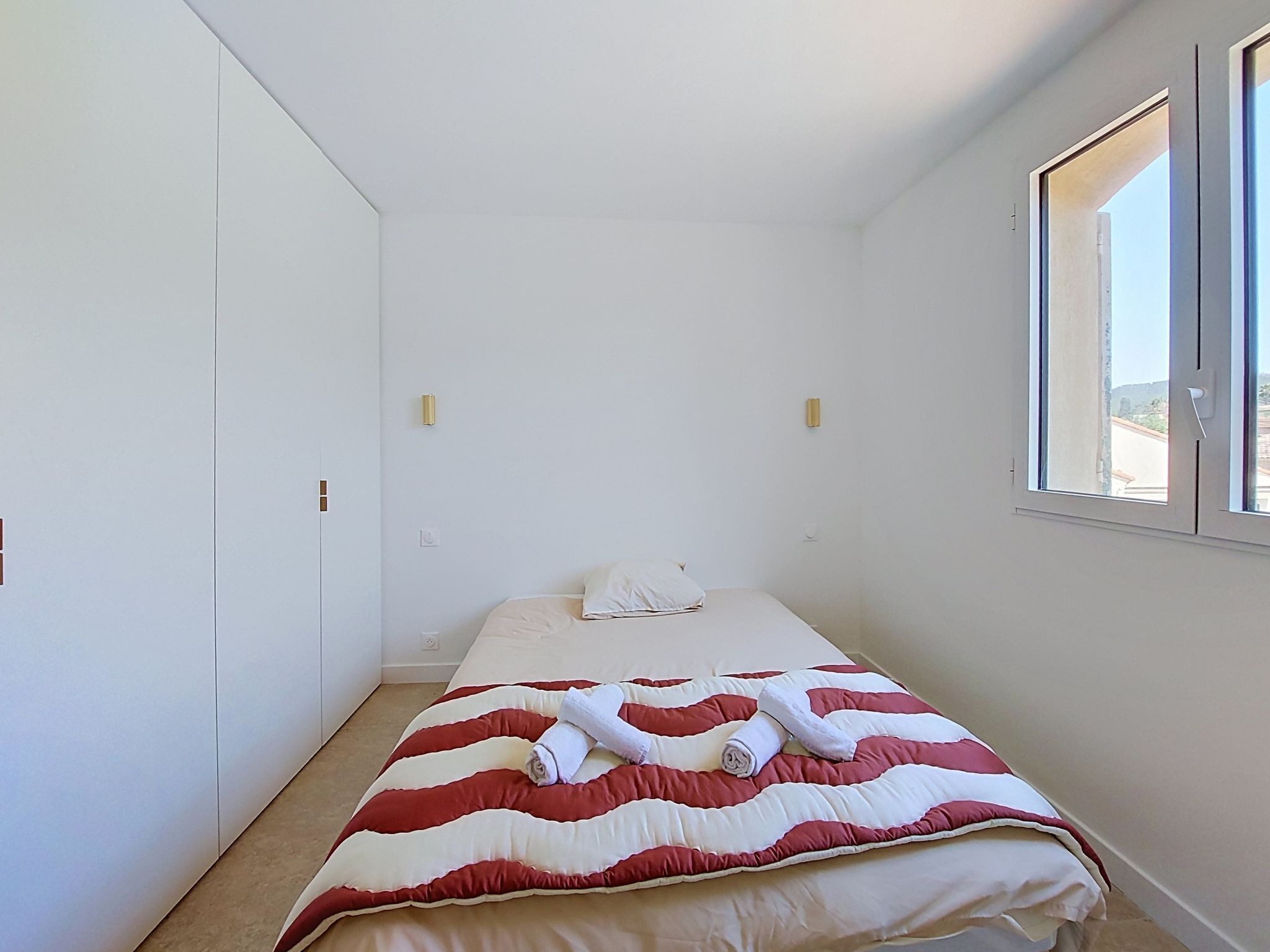 A single bed with a red and white striped bedspread and two white pillows. There is a window on the right side of the room.