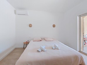 Une chambre d’hôtel avec un lit, deux lampes fixées au mur, un appareil de climatisation et une petite table sur laquelle se trouvent une bouteille et un récipient.