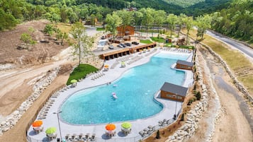 A large, curved swimming pool with lounge chairs and umbrellas, surrounded by a stone wall and a wooden building with a covered patio.