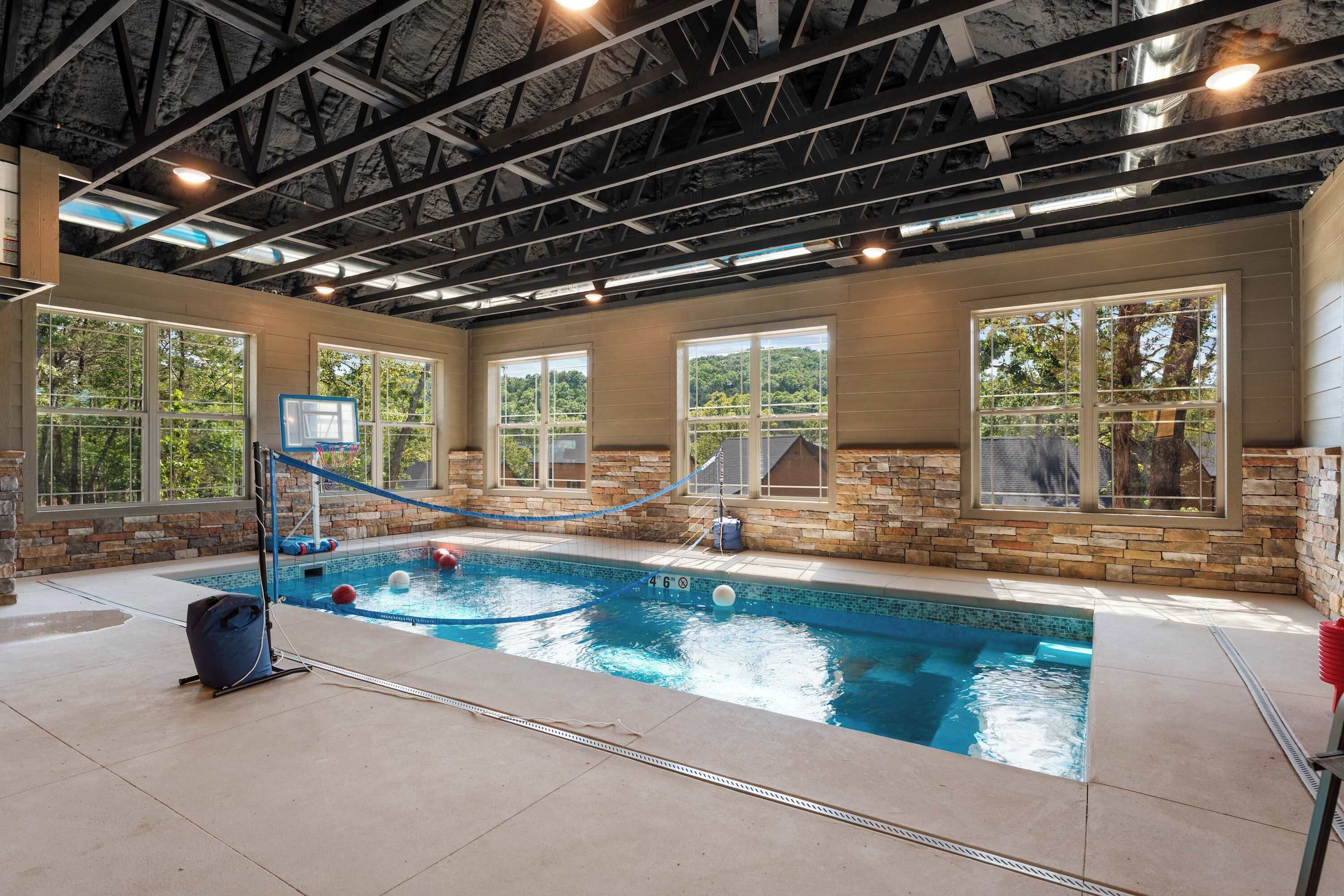 Una piscina interior con aro de baloncesto, paredes de piedra y ventanas amplias.