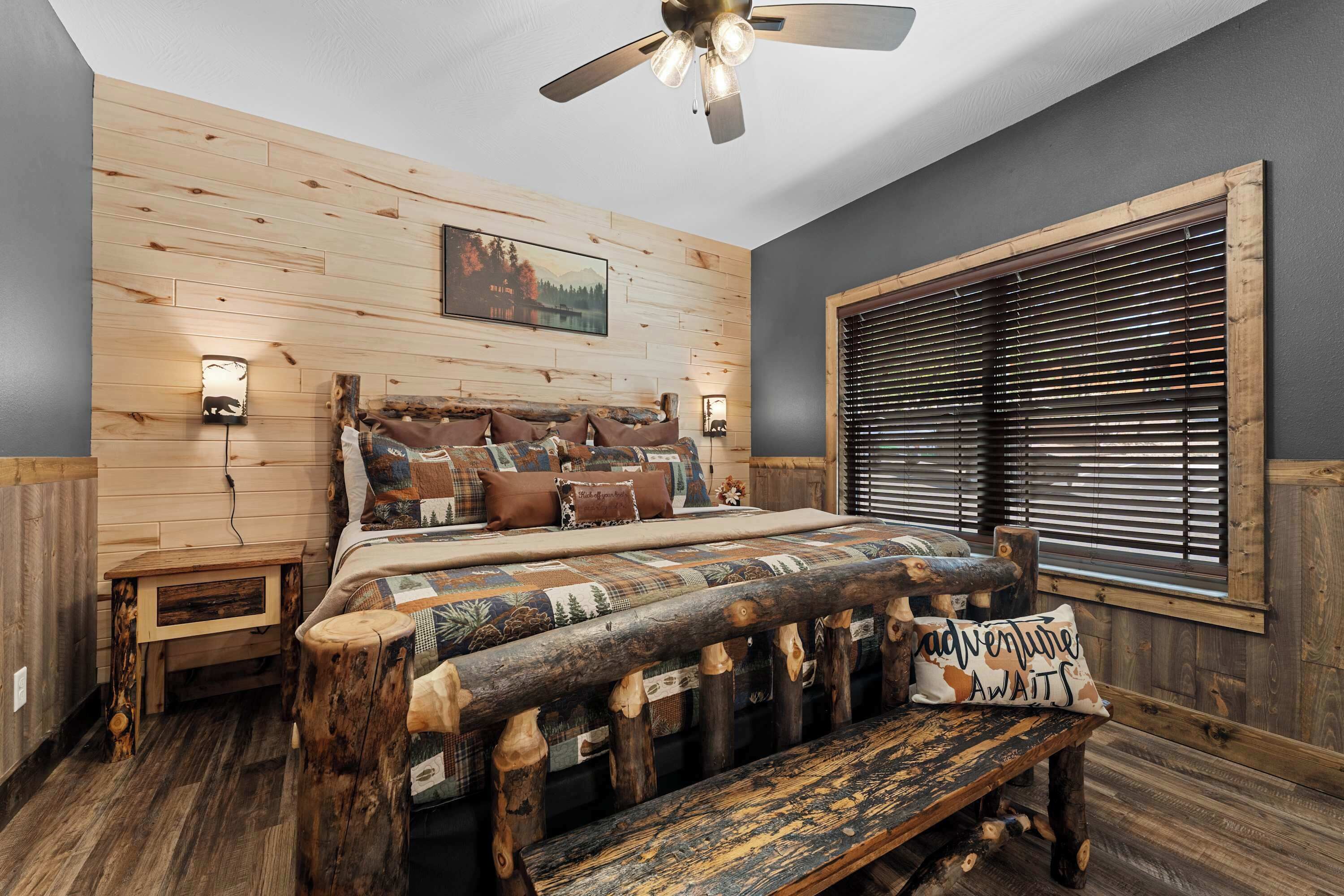 A bedroom with a wooden headboard, a bed with patterned bedding, a bench, a ceiling fan, and a window with blinds.