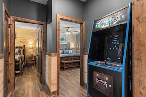 A hallway with a wooden door, a room with a bed and a ceiling fan, and an arcade machine. - New Cabin in Tall Timbers! Private Indoor Pool! (Hollister)