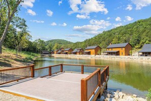 A wooden deck with a stone border leading to a row of cabins by a calm lake, surrounded by forested hills. - New Cabin in Tall Timbers! Private Indoor Pool! (Hollister)