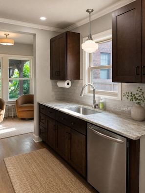 A modern kitchen with dark wood cabinets, a stainless steel dishwasher, a marble countertop, and a window with a view of greenery.