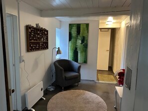 A room with a radiator, a grey armchair, a round beige rug, a floor lamp, a wall-mounted shelf with decorative items, and a large abstract painting. - Enjoy island life in a Sejerø holiday home (Sejerø)
