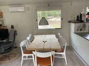 A dining area with a wooden table and white chairs, a window with a view of trees, a kitchen counter with utensils, and a TV on a stand. - Cosy little cottage in the woods at Dueodde (Nexø)
