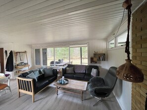 A cozy living room with a sofa, armchair, and coffee table, large windows, and a hanging light fixture. - Cosy little cottage in the woods at Dueodde (Nexø)