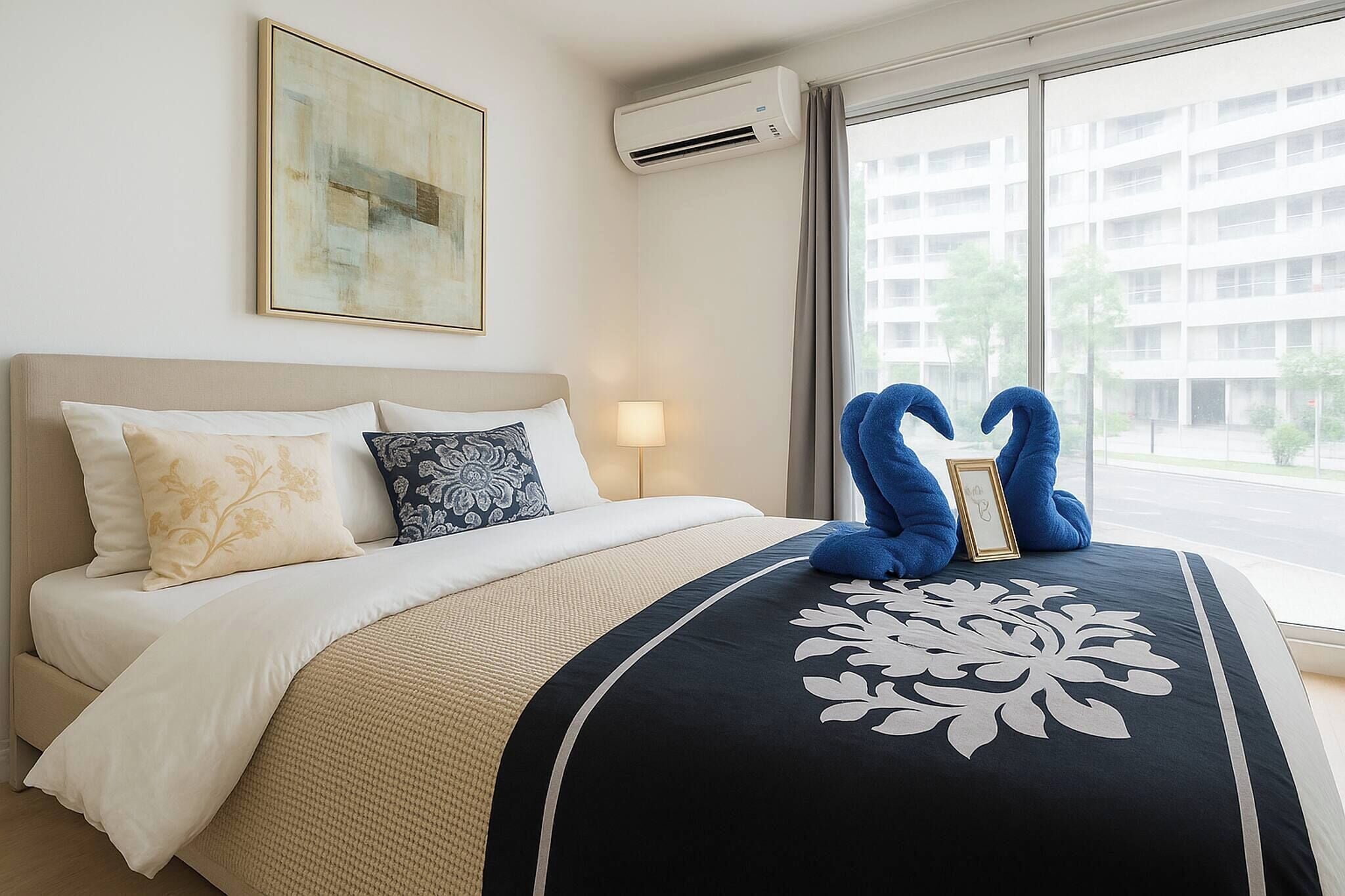 A hotel room with a bed, a wall-mounted air conditioner, a window with a view of buildings, and a framed picture on the bed.