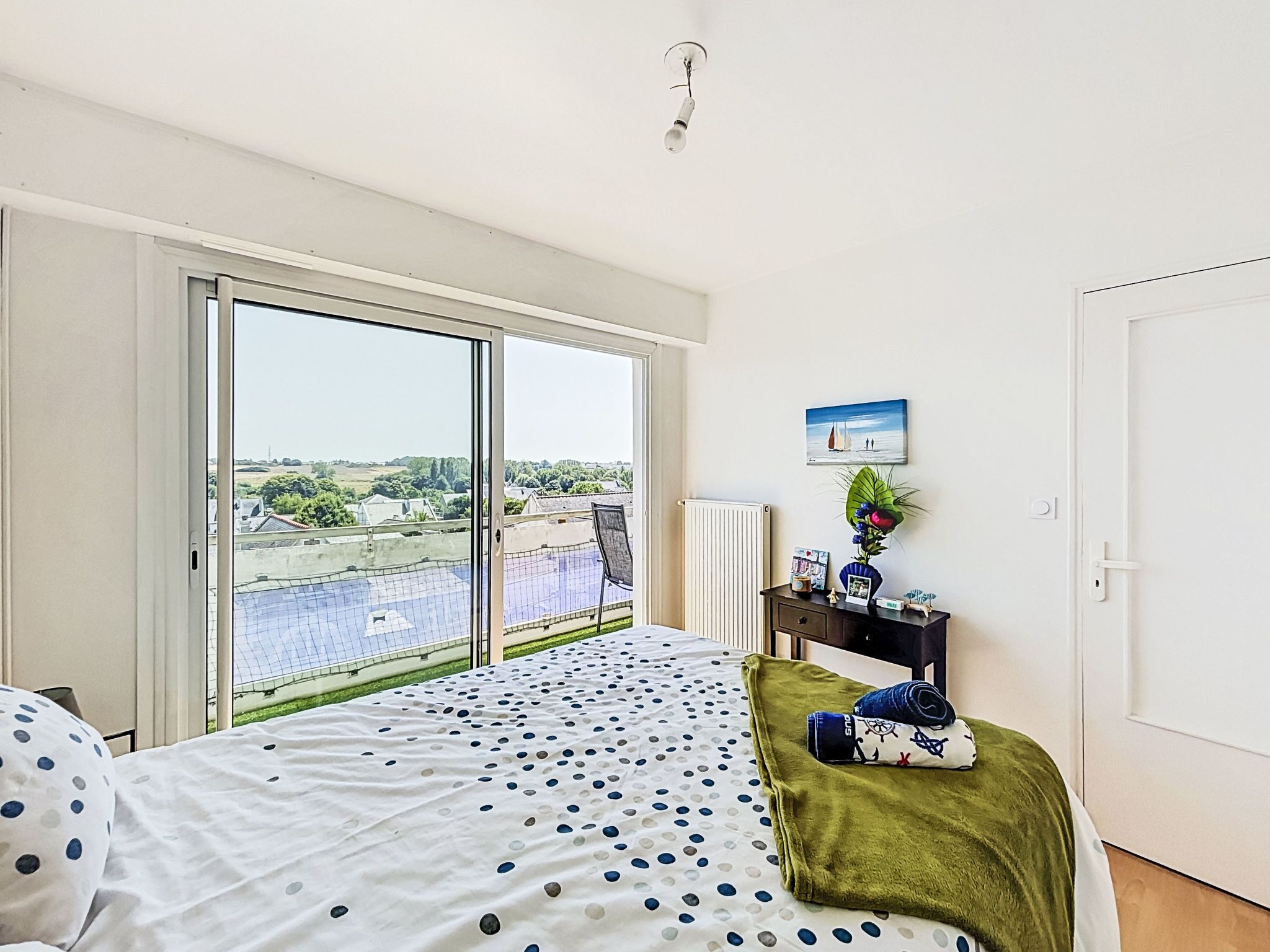 A bedroom with a large window, a bed with a polka dot bedspread, a green throw blanket, a nightstand with a vase of flowers, and a painting of a sailboat on the wall.