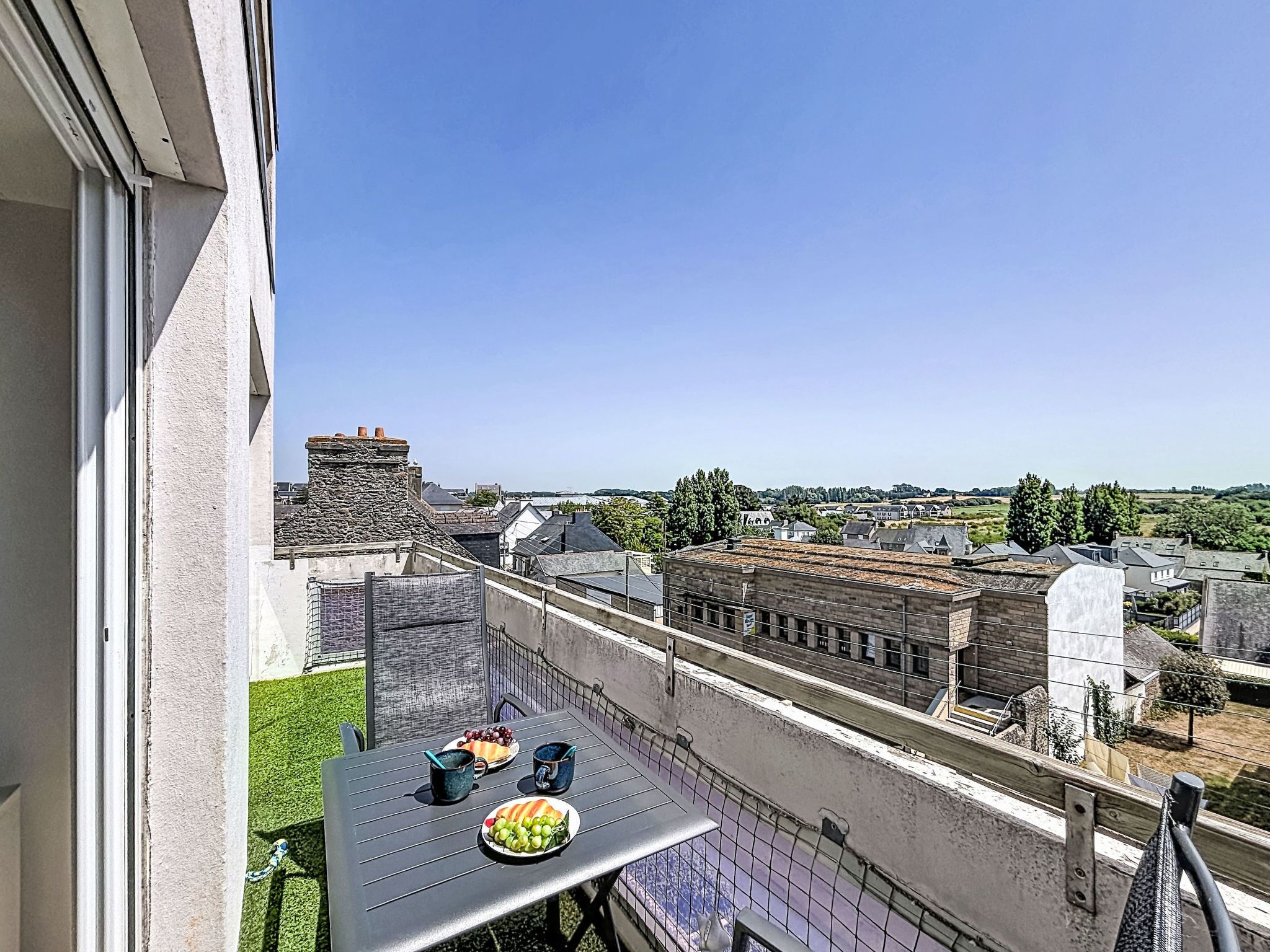 Un balcon avec une table dressée pour deux personnes, donnant sur un quartier résidentiel avec des maisons et un ciel dégagé.