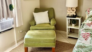 Ein Zimmer mit grünem Sessel, weißem Kissen, kleinem Tisch mit Lampe, einem gerahmten Bild an der Wand und einem Bett mit blumigem Überwurf.
