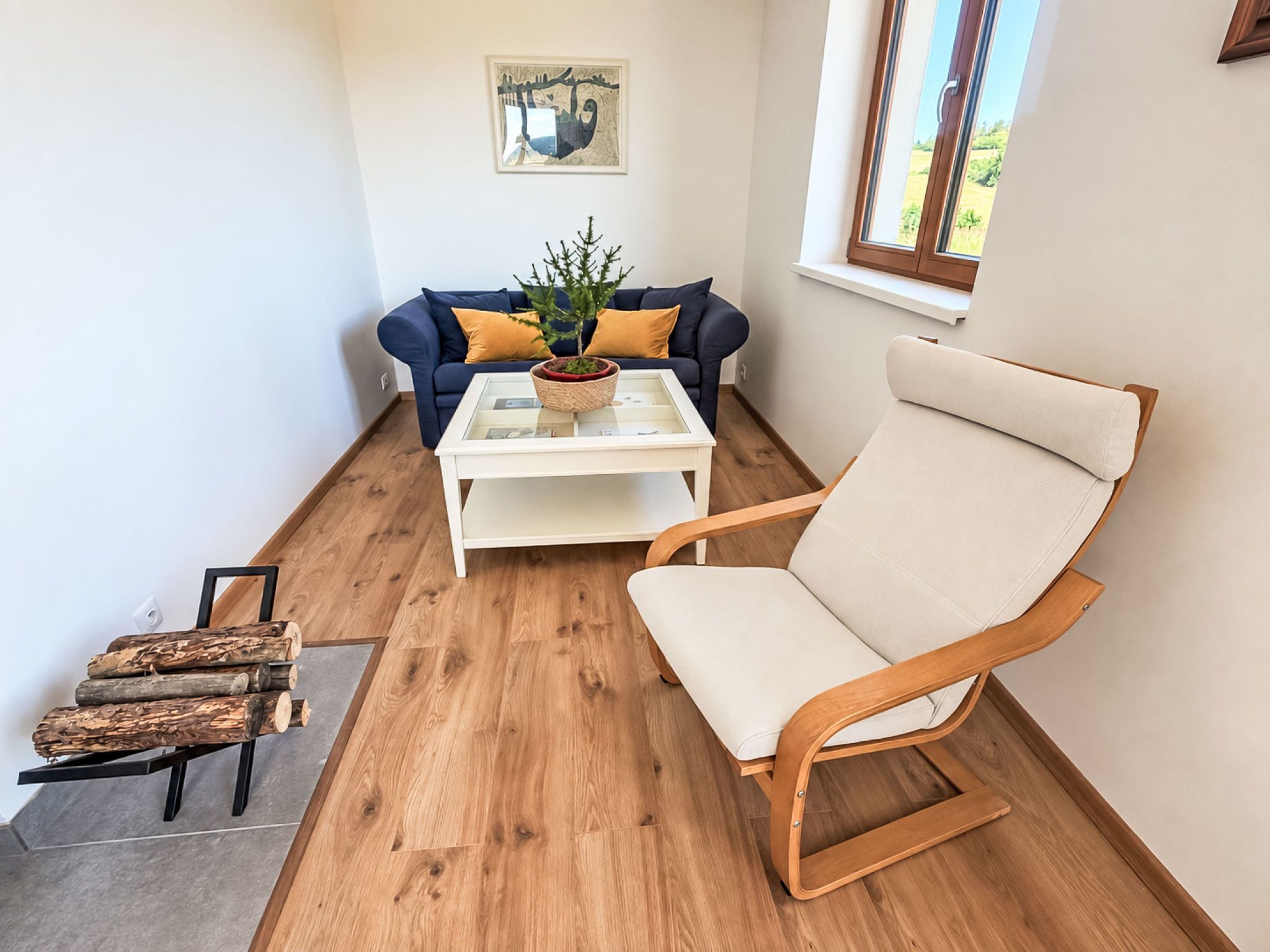 A room with a blue sofa, a white coffee table, a wooden chair, and a window with a view of greenery.