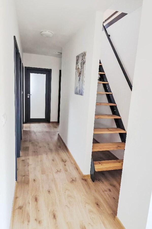 Un pasillo con piso de madera, una puerta negra al final y una escalera con peldaños de madera.