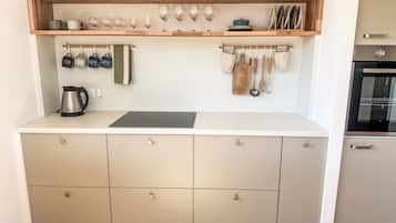 A kitchen with a wooden shelf holding glasses and a kettle, a countertop with a cutting board, and a hanging pot rack.