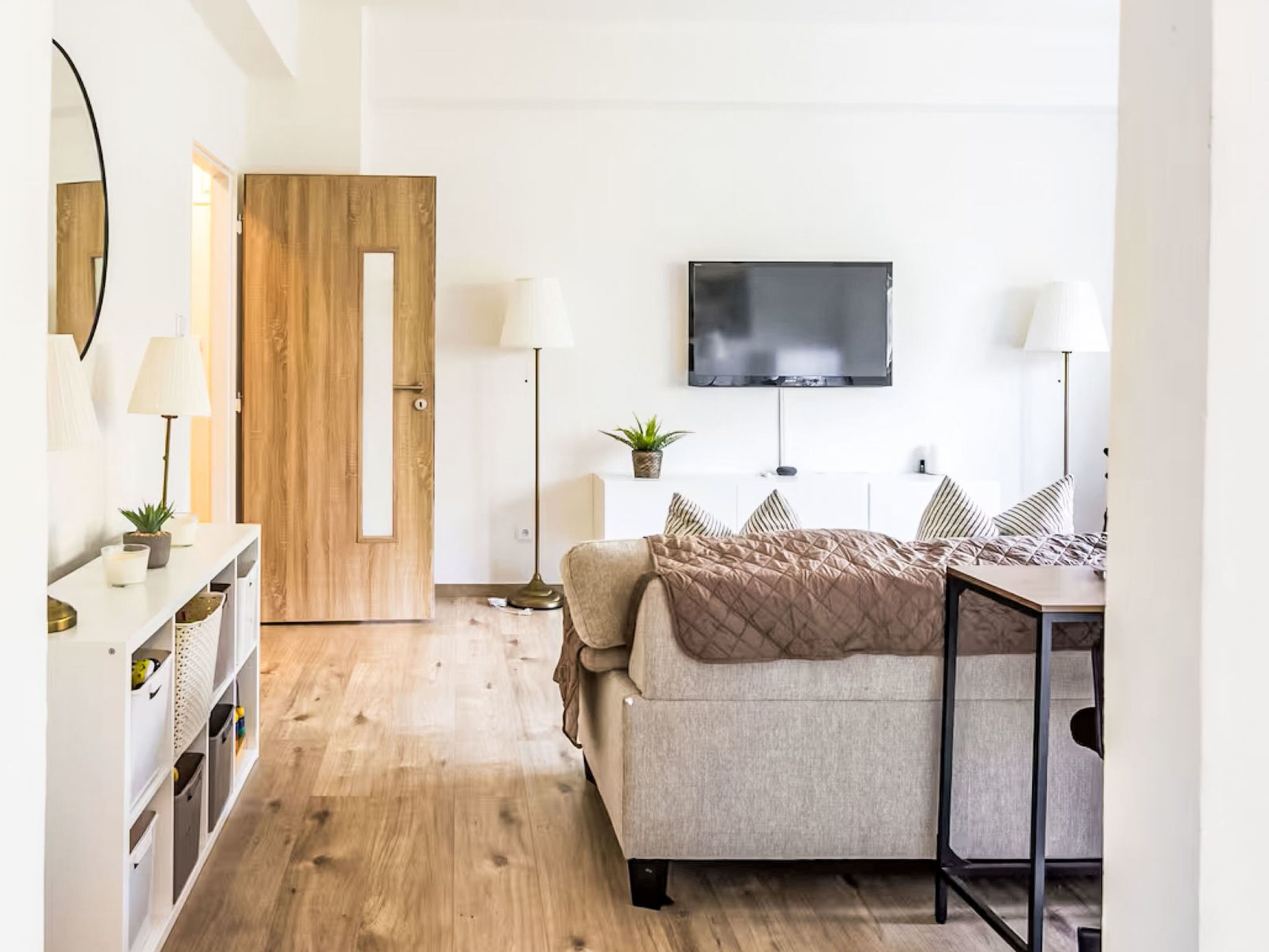 Quarto com cama, luminárias de cabeceira, televisão, porta de madeira e mesa de consola branca com objetos decorativos.