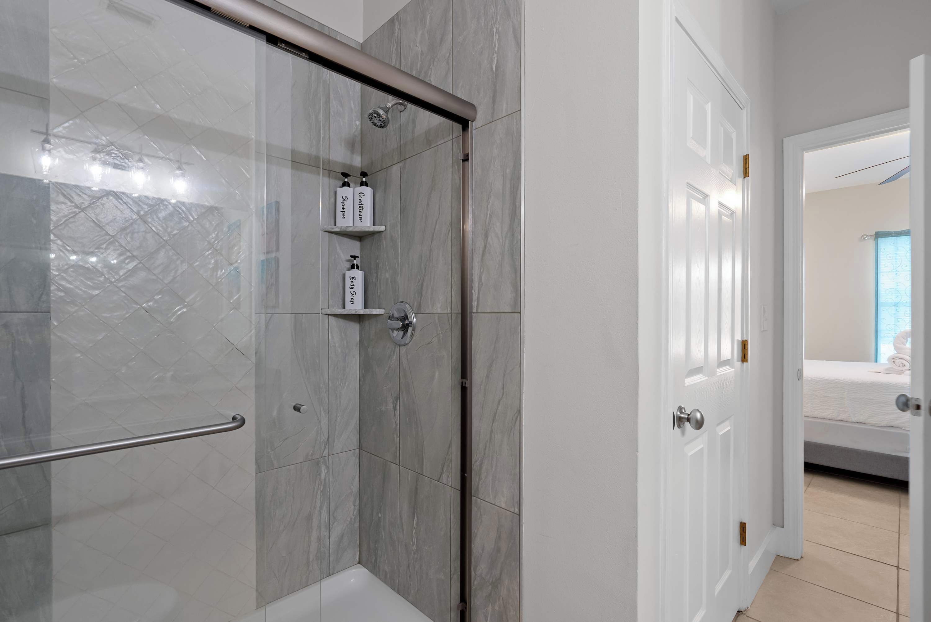 A bathroom with a glass shower enclosure, a white bathtub, and a door leading to a bedroom.