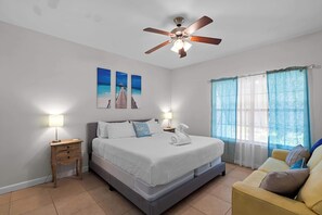 A bedroom with a bed, a nightstand, a ceiling fan, a window with curtains, and artwork on the wall.