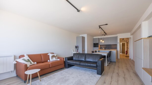 A modern living room with a sofa, a coffee table, and a rug. There is a kitchen area with a dining table and chairs. - 8th-floor apartment with sea view (Middelkerke)