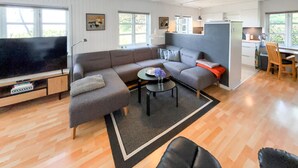 Ein modernes Wohnzimmer mit einem großen Flachbildfernseher, grauen Sofas, einem Couchtisch und einer Küchenzeile mit Kühlschrank und Holzstühlen.