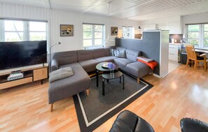 Ein modernes Wohnzimmer mit einem großen Flachbildfernseher, grauen Sofas, einem Couchtisch und einer Küchenzeile mit Kühlschrank und Holzstühlen.