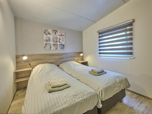 A small, single-room accommodation with two beds, a wooden headboard, a wall-mounted picture, and a window with blinds. - Beach lodge on the beach (Callantsoog)
