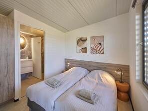 A modern bedroom with a bed, two bedside tables, a nightstand, a wall-mounted lamp, and a window with blinds. - Luxury beach lodge by the beach. (Callantsoog)