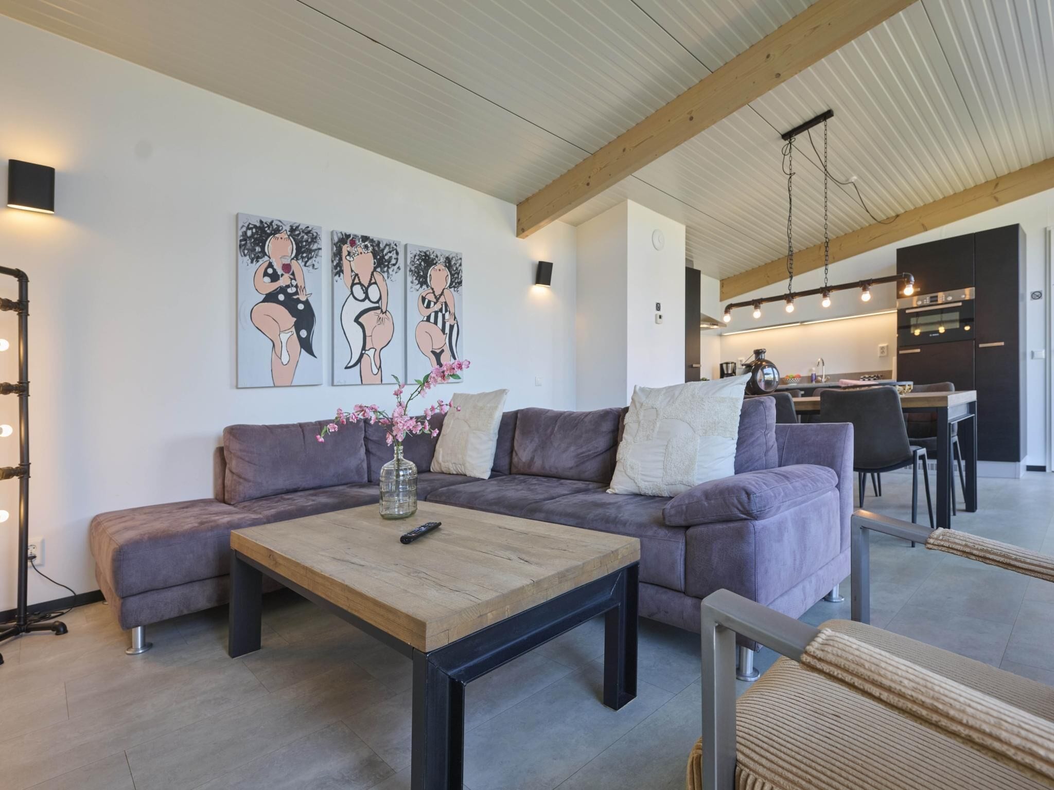 Ein modernes Wohnzimmer mit einer Couch, einem Couchtisch und einer Blumenvase. Im Hintergrund ist eine Küche mit einem Essbereich zu sehen.