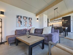 Ein modernes Wohnzimmer mit einer Couch, einem Couchtisch und einer Blumenvase. Im Hintergrund ist eine Küche mit einem Essbereich zu sehen.