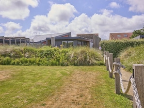 A grassy area with a path leading to a modern building with large windows and a sloped roof. - Holiday home 350 meters from the beach (Callantsoog)