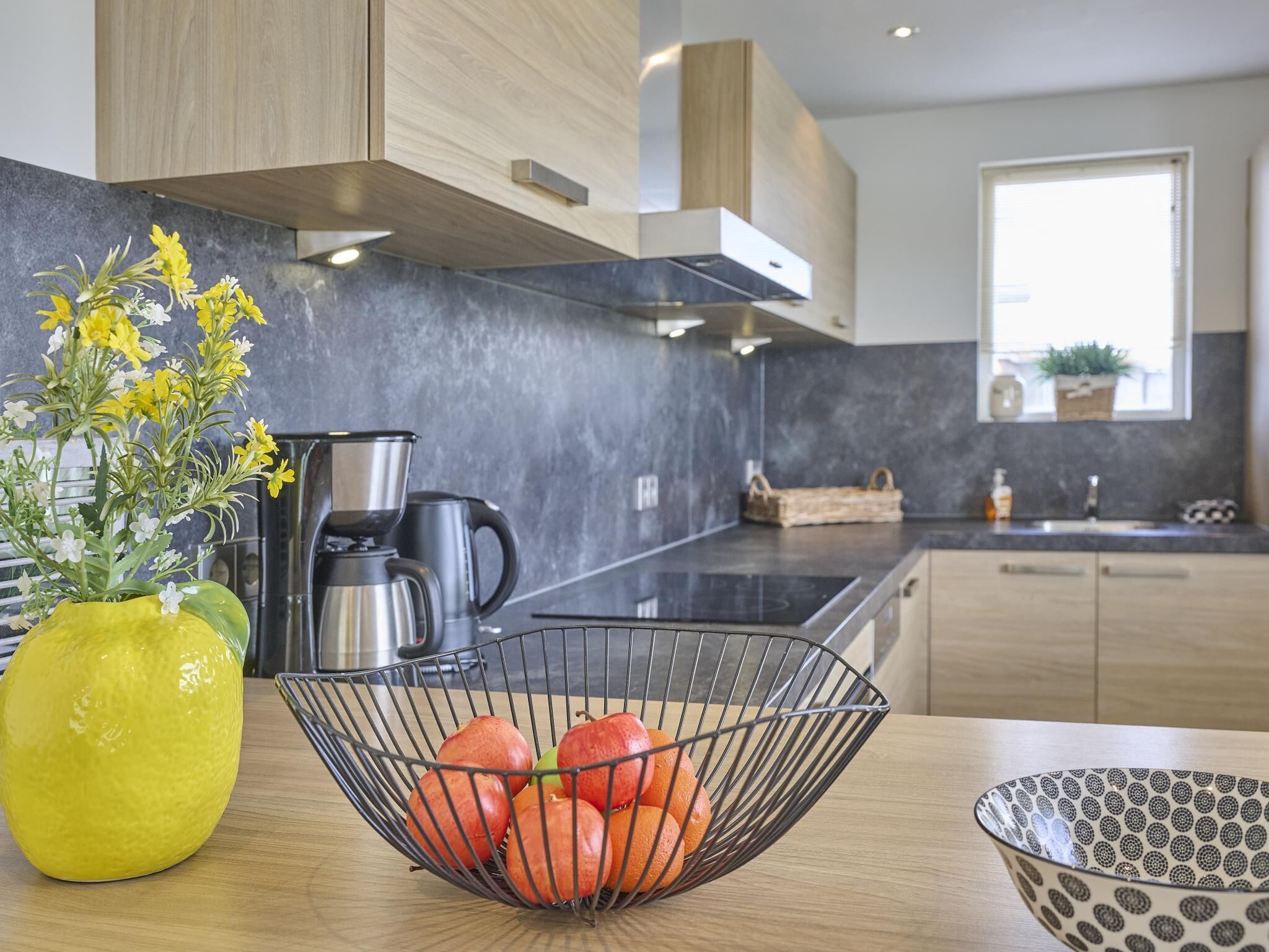 A modern kitchen with a black countertop, a yellow vase with flowers, and a metal fruit basket.