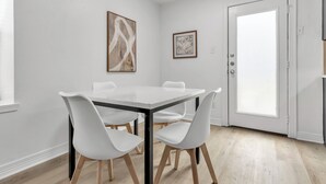 Un comedor con una mesa y sillas blancas, una puerta con panel de vidrio y dos cuadros en la pared.