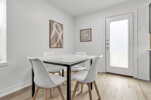 Een eethoek met een witte tafel en stoelen, een deur met een ruitpaneel, en twee ingelijste kunstwerken aan de muur.