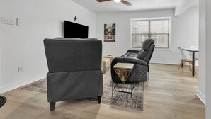 Una sala de estar moderna con un ventilador de techo, un sofá negro, una mesita auxiliar y una zona de comedor con una mesa y sillas blancas.