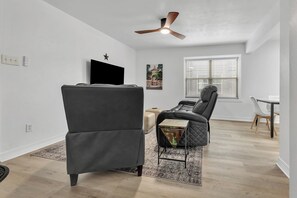 Een moderne woonkamer met een plafondventilator, een zwarte bank, een klein bijzettafeltje en een eethoek met een witte tafel en stoelen.
