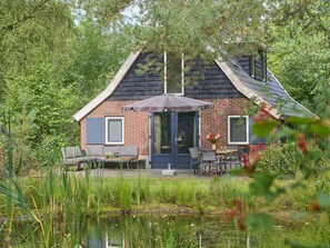 A house with a brick exterior, a covered patio with outdoor furniture, and a pond in the foreground. - Nice 8-person house in a child-friendly park. (Otterlo)