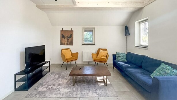 Een woonkamer met een televisie, twee gele fauteuils, een blauwe bank, een salontafel en kunstwerken aan de muur.