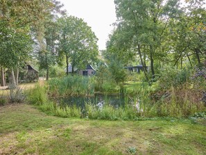 A serene lakeside setting with a small house and a larger building in the background, surrounded by lush greenery and tall grasses. - Nice 8-person house in a child-friendly park. (Otterlo)