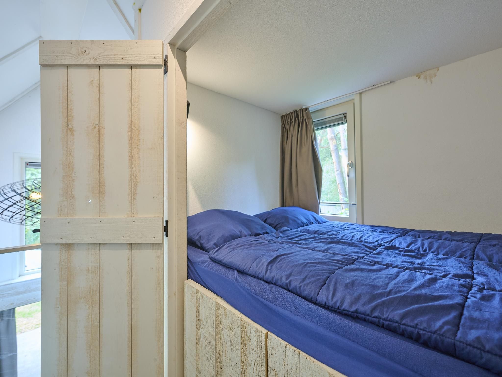 Un lit simple avec un matelas bleu dans une pièce aux murs blancs et dotée d’une fenêtre avec des rideaux.