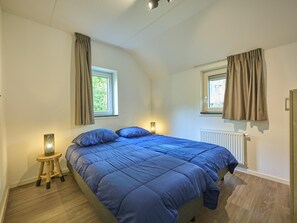 Une chambre à coucher avec un lit, deux fenêtres avec des rideaux, un radiateur et une petite table avec une lampe.