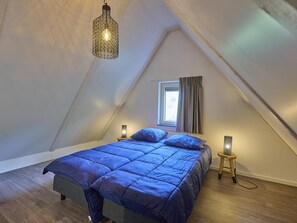 A bedroom with a sloped ceiling, a bed with blue bedding, two bedside lamps, and a window with curtains. - Nice 8-person house in a child-friendly park. (Otterlo)