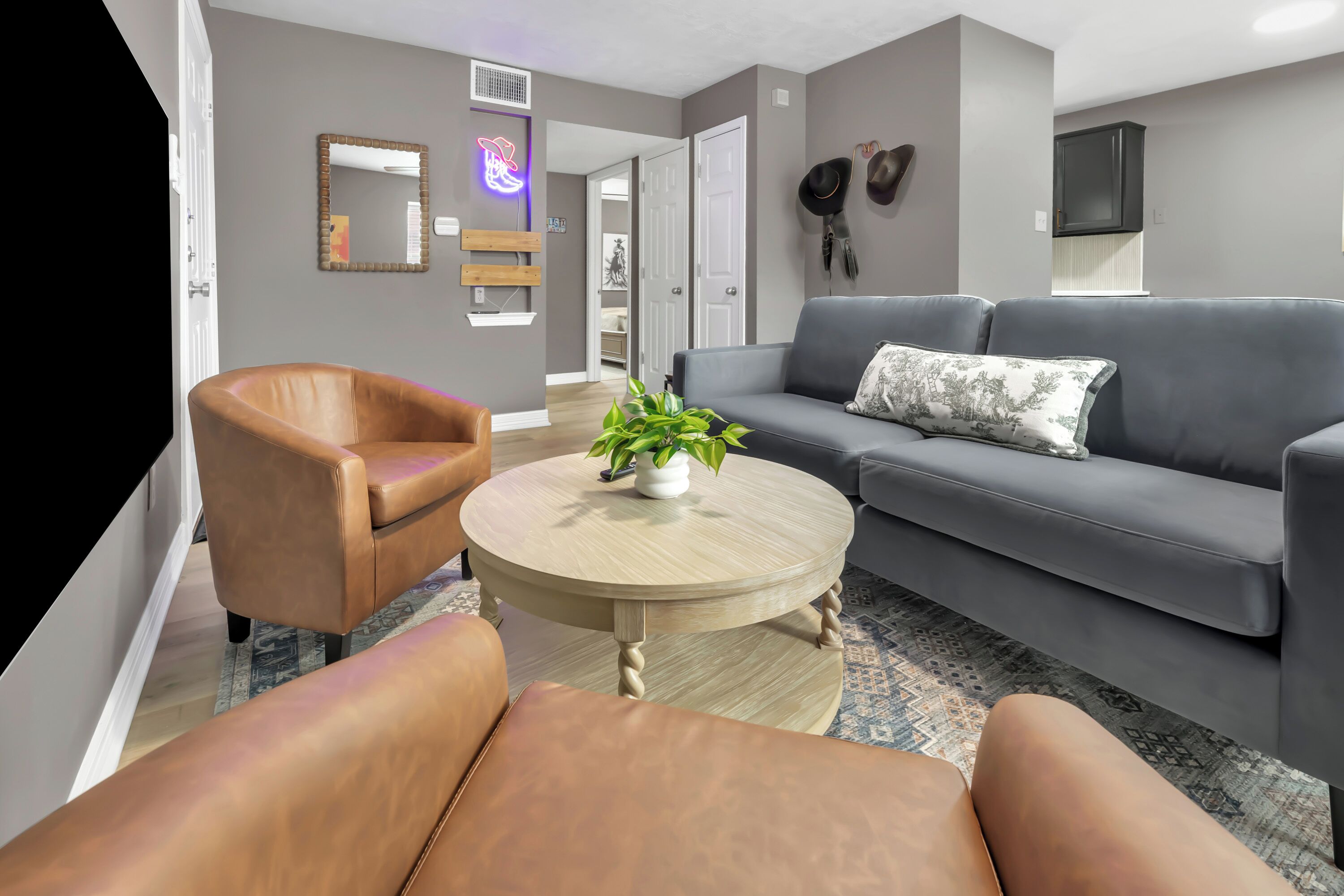 A living room with a grey sofa, a brown leather chair, a round wooden coffee table with a plant, and a mirror with a neon light.