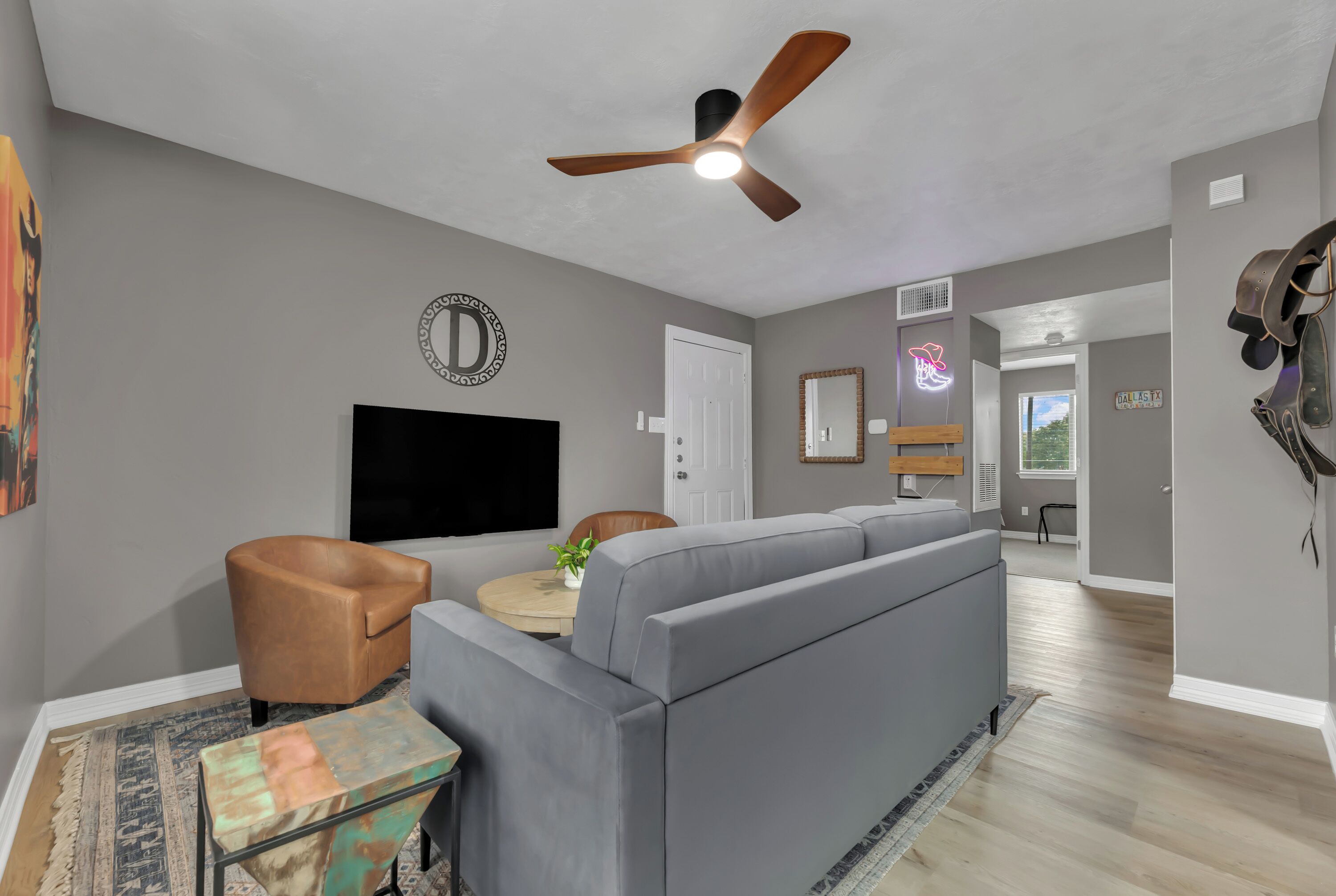 A modern living room with a grey sofa, a ceiling fan, a flat-screen TV, and a decorative wall piece.