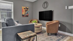 A living room with a grey sofa, a brown leather chair, a round coffee table, a flat-screen TV mounted on the wall, and a patterned rug on the floor. - Dallas: Walk to Texas A&M Campus, Comfy King Beds (Bryan)