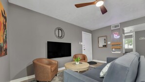 A living room with a grey sofa, a brown leather chair, a round coffee table with a plant, a flat-screen TV mounted on the wall, and a ceiling fan. - Dallas: Walk to Texas A&M Campus, Comfy King Beds (Bryan)
