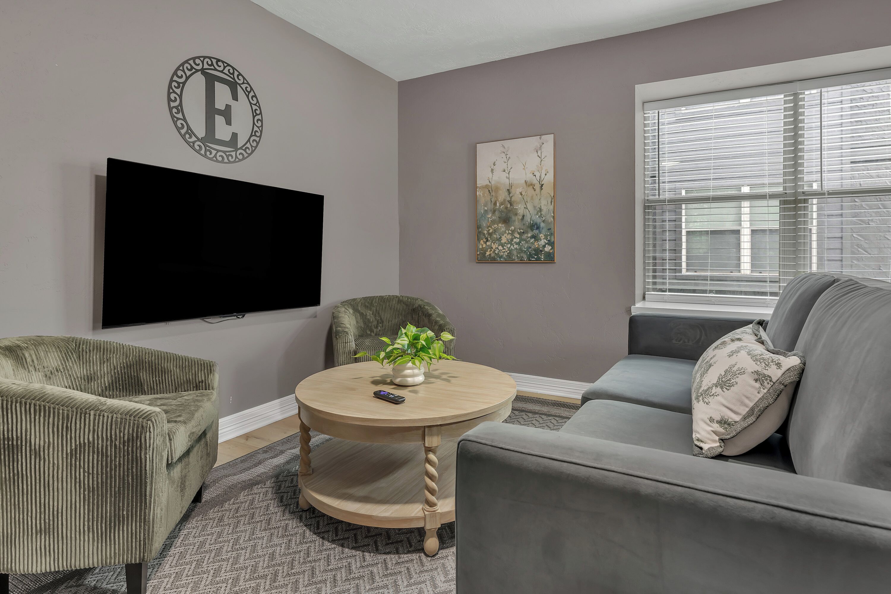 A living room with a flat-screen TV, a round wooden table, a grey sofa, and a chair.